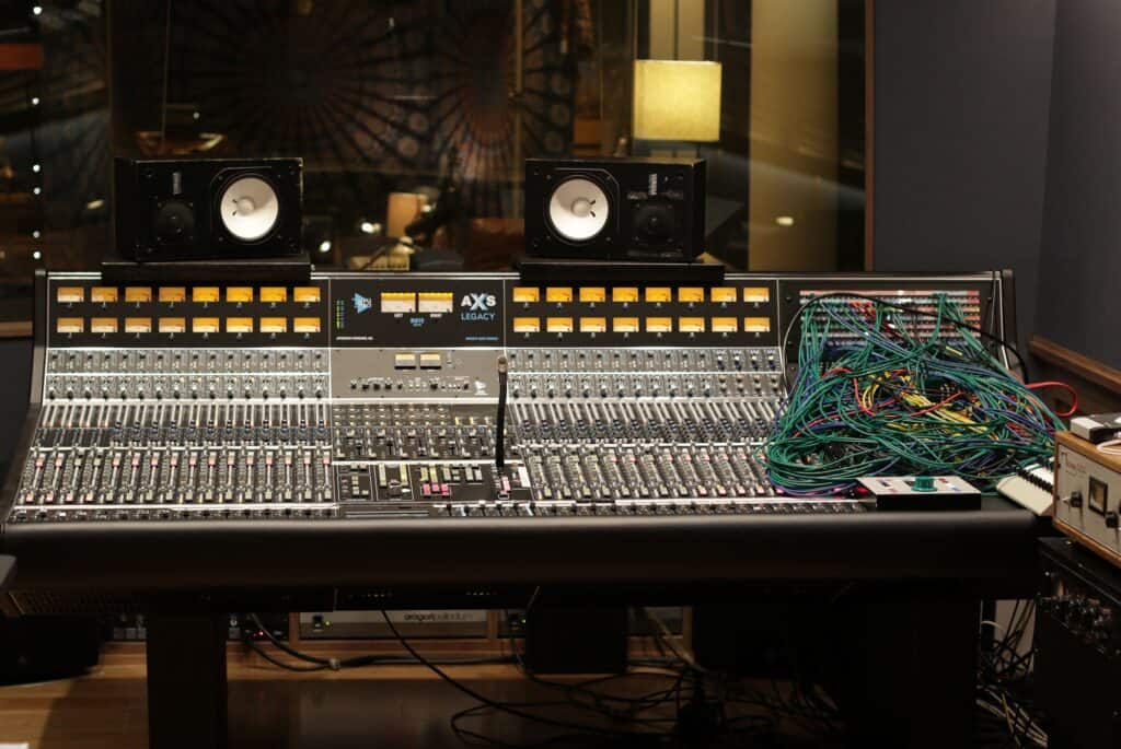 API 32-channel AXS console in commercial studio with patchbay on right side, cables overflowing, looking into live room with yellow lamp